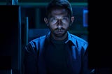 A dimly lit computer lab with a lone computer screen casting a blue glow on the face of a nervous-looking brown grad student.