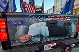 The tailgate of a pick-up truck. Text across the top reads Trump 2024, with a ‘Let’s Go Brandon’ bumper sticker to the left. Below is an image of President Biden hogtied in the bed of the truck.