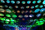 Groups of people gathered together to sing and dance and do human things, with umbrellas lining the “ceiling” of the clearing in the trees, and lights coming from the far end.