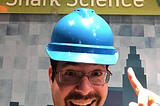 A man (Dr David Shiffman) with a goatee and glasses wearing a hard hat and smiling while pointing at a sign above his head that reads “Shark Science”