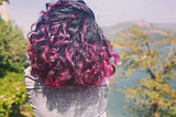 A woman with curly pink hair admiring the lake facing the camera with her back.