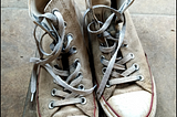 A pair of sneakers, Converse high tops, dirty and worn, rest on a tile floor.