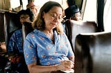 Mulher branca, cabelos castanhos claros, está sentada no ônibus de viagem e está escrevendo em um papel. Ela veste uma camisa azul com estampa de flores na cor rosa.