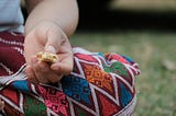 close-up on a person sitting on the ground wearing a brightly colored skirt pulled over the knee, with hand resting on the leg. In the hand is a small, remaining bite of a crispy, protein bar.