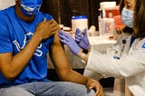 A young man in a blue shirt and blue mask receiving a Covid-19 vaccination in his left arm by a gloved and surgically masked woman in a white lab coat