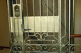 A color photograph an old elevator, resembling a cage. The metal is golden in appearance, with metal bars in a decorative pattern. There is a protective screen, also golden in color, and well as a large metal plate. There is also a handle and what a device with two small buttons. At the top is a sign that appears to be indicating that this elevator can carry 4 persons.