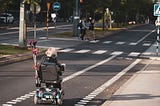 A person on a mobility scooter is riding down the empty street on a dedicated lane towards a crossroads. Two people are about to cross the street on a pedestrian crossing
