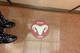 A circular “stand here” sticker on the floor of an elevator, with a Ram’s head mascot in the middle. Two feet in Oxfords.