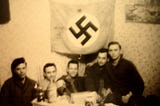 295th men (Rogers, Deragio, Franklin, Sage, and Jack Hope) sitting at a kitchen table with Jack’s captured Nazi flag.
