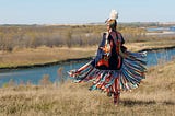 First Nations Women performing a Fancy Shawl Dance in a grass field with a river background