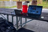 a park bench and table with a laptop, giant red jug with a straw, a seat cushion, backpack and binders with water and a fountain in the background, surrounded by grass and trees