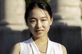 Portrait of AfroLA’s first community engagement fellow, Heather Wang. She’s smiling at the camera in a white top with stone columns in the background.