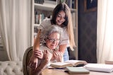 Grandmother sitting at table reading books, granddaughter has her hands wrapped around grandmother’s shoulders in a hug