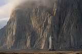 A gleaming modern rocket stands dwarfed at the base of an immense granite cliff that fills the frame, symbolizing human ambition and capital confronting the immovable laws of physics.