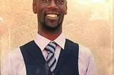 photograph of smiling Black man in a light pink shirt, striped tie, and charcoal suit vest