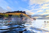 A dark lagoon merges seamlessly with the ocean beyond under a blue sky and a small hut is visible with a rocky top
