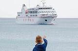 Young boy dressed in blue waving goodbye to a cruise ship.