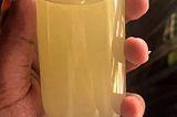 A woman’s hand holding a small glass spray bottle that has yellow liquid in it that looks like lemonade. A plant is in the background.