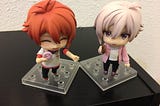 two Nendoroid figures, one with red hair and a cheerful expression, and one with white hair and blushing stand side by side.