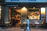 A woman carrying a white plastic bag walks by Paris restaurant JeanneA at night. The restaurant has sidewalk tables.