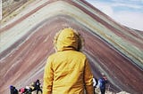 A person in a mustard yellow fur-lined hood stands facing Peru’s Rainbow Mountain, with its vibrant natural stripes of red, green, and gold layered across the peak under a partly cloudy sky. Other hikers rest and take photos in the background.