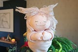 Photo of a pink Christmas tree fairy. She has the most beautiful smile and her hands are clasped together in front of her. Her hair is made of pink cotton threads and it is sticking out at funny angles. The blurred background shows green foliage, a poinsettia, a candle and other Christmas decorations.