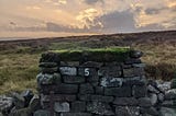 A moss-covered dry stone wall with the number 5 painted on it, a cloudy sunset in the background.