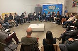 A group of 30 people sit in a circle. There is a low white square table in the center of the room and a large artwork with a black and white drawing against a bright blue background.