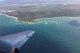 A plane wing out a plane window flying over the turquoise sea and white beaches of an island.