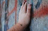 A close-up of a person’s hand resting gently on a worn, graffiti-covered concrete wall. The wall is cracked and layered with faded red and blue spray paint. The hand is steady and open, suggesting quiet presence or reflection.
