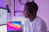 A Black man wearing headphones, in front of two monitors on a desk.