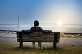 Person sitting on a bench in front of a lake looking at a suspension bridge with the sun rising