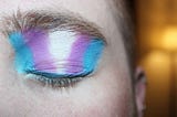 A close-up of a closed eye with the trans flag colours—blue, pink and white—painted on it with eyeshadow.