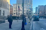 Two people on a sidewalk in front of a courthouse bulding; a bus with the # 4 approaching.