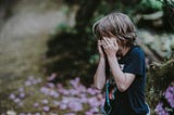 Boy blue shirt hands over eyes in garden purple flowers