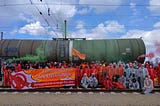 Aktivisti blockieren das Raffineriegleis vor einem Tankerwagen. Verkleidet in orangen und weissen Anzügen mit einem Transpi worauf „Klimazerstörin OMV enteignen! Klimagerechte Energie für alle!’’