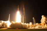 A rocket launch at night. The glow from the rocket illuminates the launchpad and smoke surrounding it as it begins to fly.