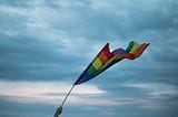 Rainbow flag held in the wind with grey clouds in the background.