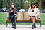 Two women sitting on a park bench paying no attention to each other