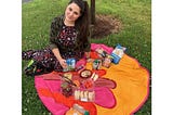 Brunette millennial in a dress sits on a round towel covered in snacks sometime in 2019.