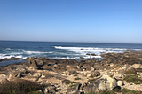Blue sky, deep blue sea with white water and waves crashing against yellow and brown rocks on shore.