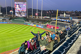 People from the Monarch team and other Biocuration Conference attendees look back towards the camera while siting in the baseball stadium with the field behind them.