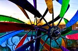 A closeup of a windmill with many colors in front of a sunset and a fence covered in graffiti writing something indeciferable.