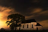 Under a dark starry sky, a white concrete haunted house with a black roof rests next to a tree.