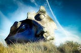Blue Sky photo of a broken mask sculpture in a grassy field.