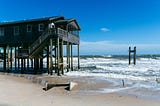 Photo of a home on stilts in the water next to a few pilings in the water from a previous home that was washed away