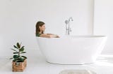 A woman wearing a shirt sits in a white bathtub, staring blankly ahead. There is a plant in a wooden basket near the tub.