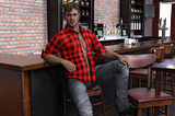 Man in unbuttoned shirt leaning against a bar.