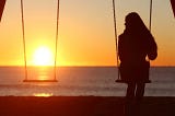 A girl sitting alone on a swing at the beach, gazing at the empty seat beside her, feeling lonely.