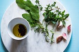 Several plants & leaves lie next to a cup filled with oil. All are resting on a circular white marble stone on a blue table